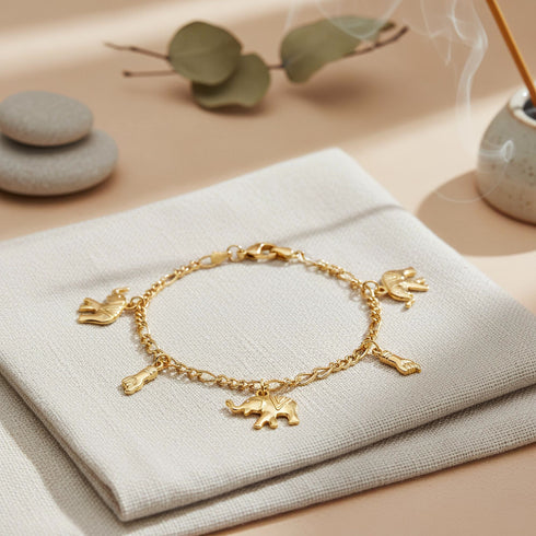14k gold-filled bracelet adorned with elephant and Figa hand charms rests on beige cloth, surrounded by stones, leaves, and incense.